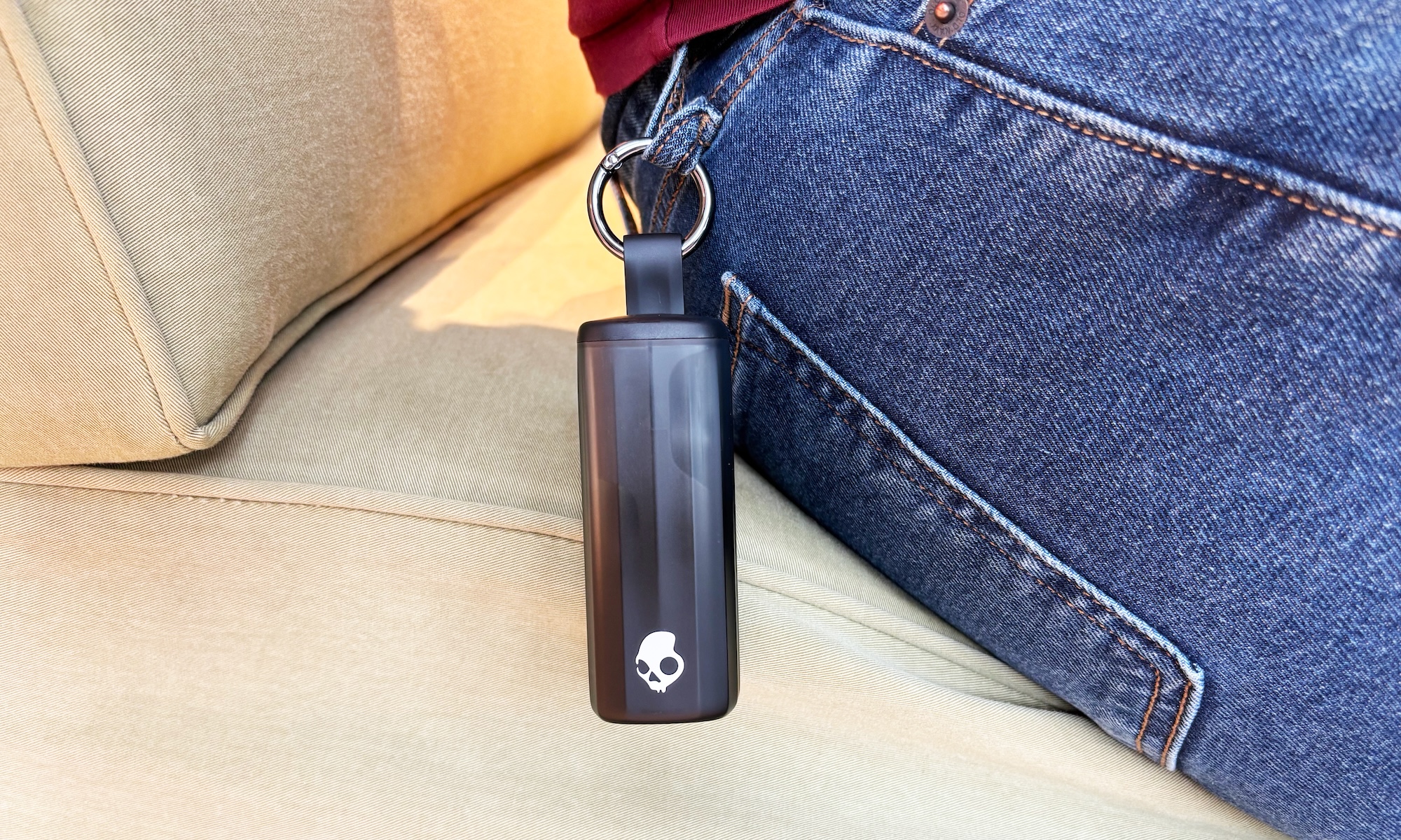 The black tube-like charging case for the Skullcandy Method 360 ANC earbuds is shown hooked onto a man's blue jeans while he sits on a light brown couch. The case is hooked to the jeans' belt loop via its built-in clip.