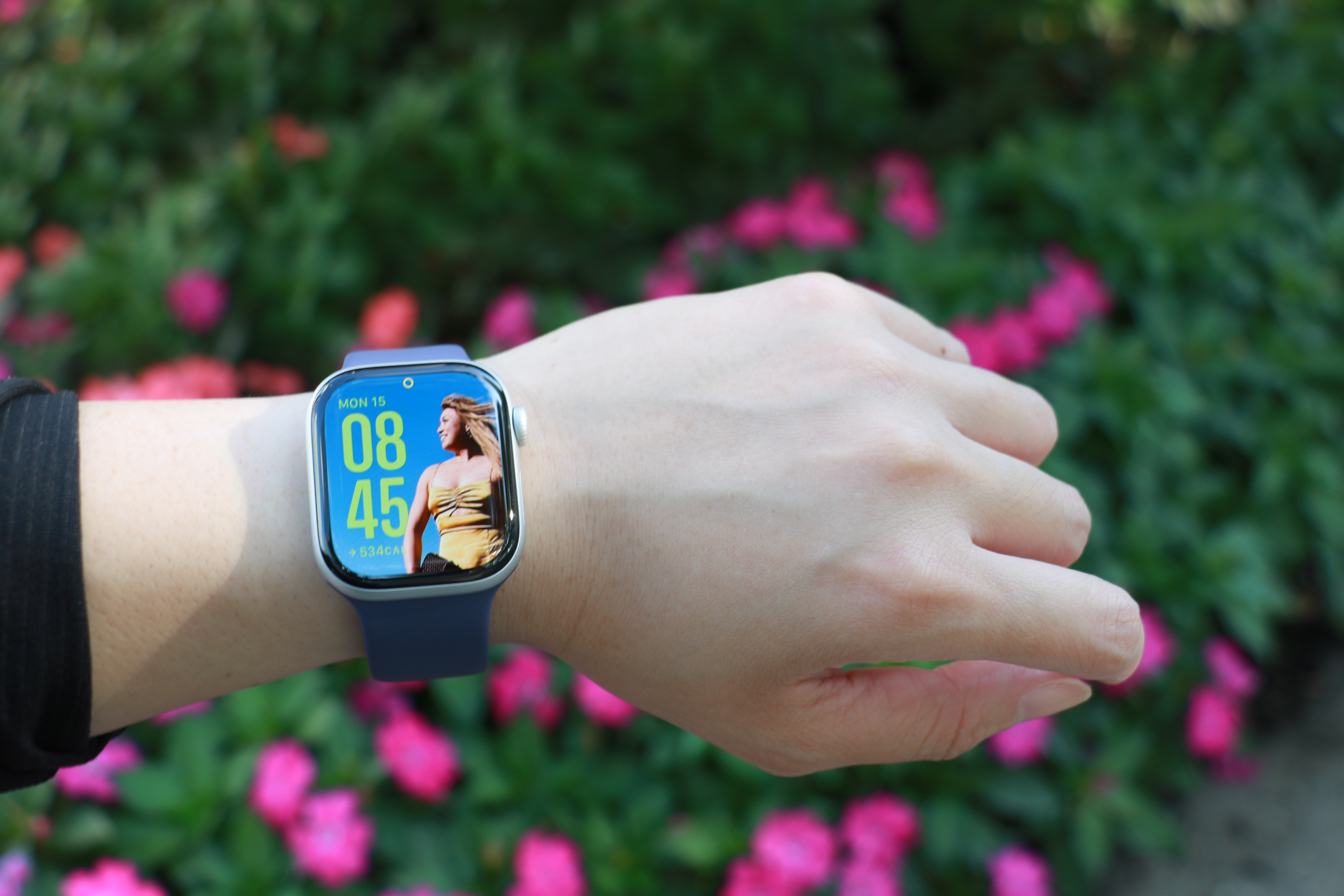 The Apple Watch Series 11 on a wrist held out in mid air in front of a bush with some pink flowers.