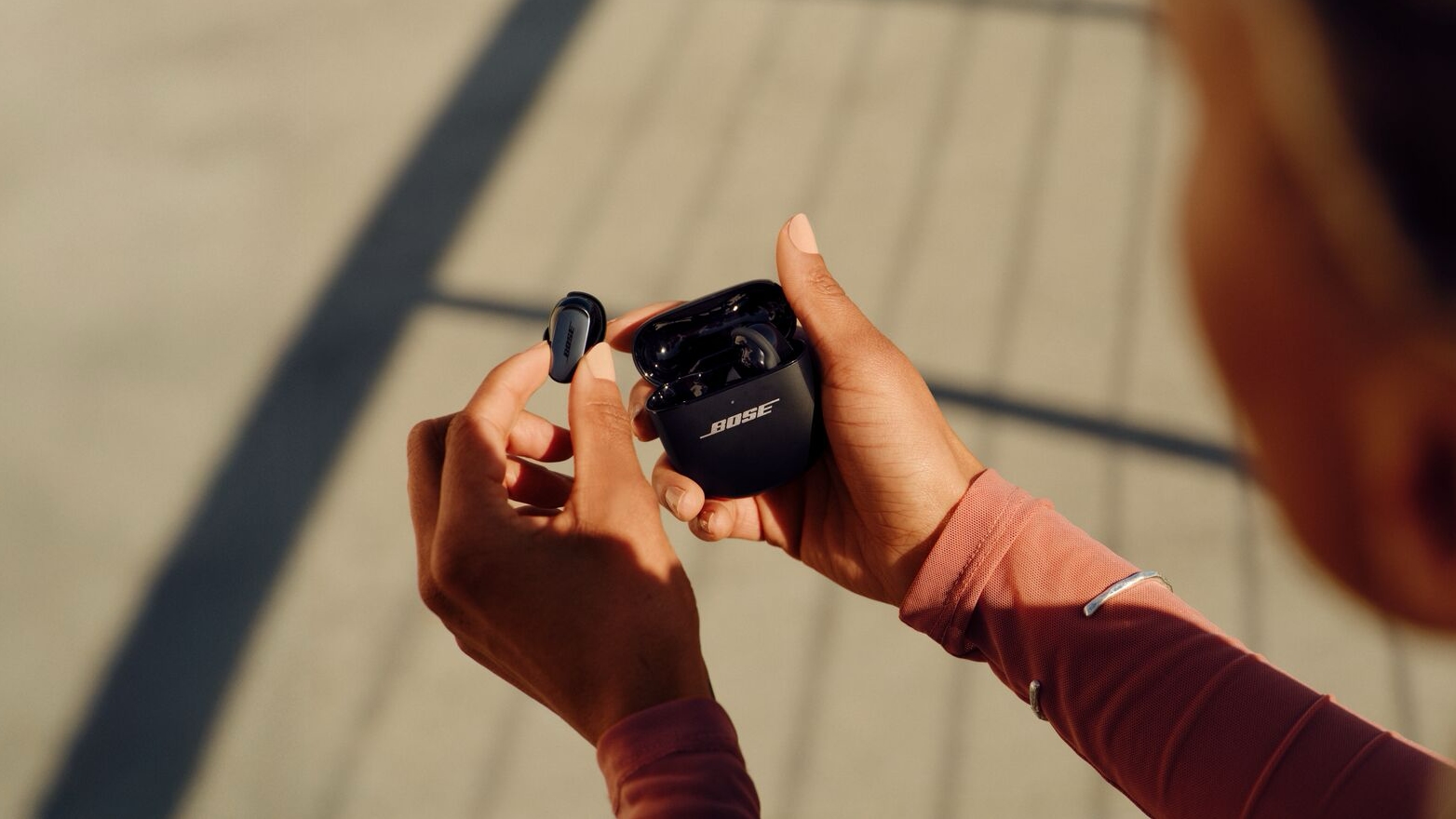 Over-the-shoulder view of someone holding the Bose QC Ultra Earbuds and their charging case. One bud is out of the case.