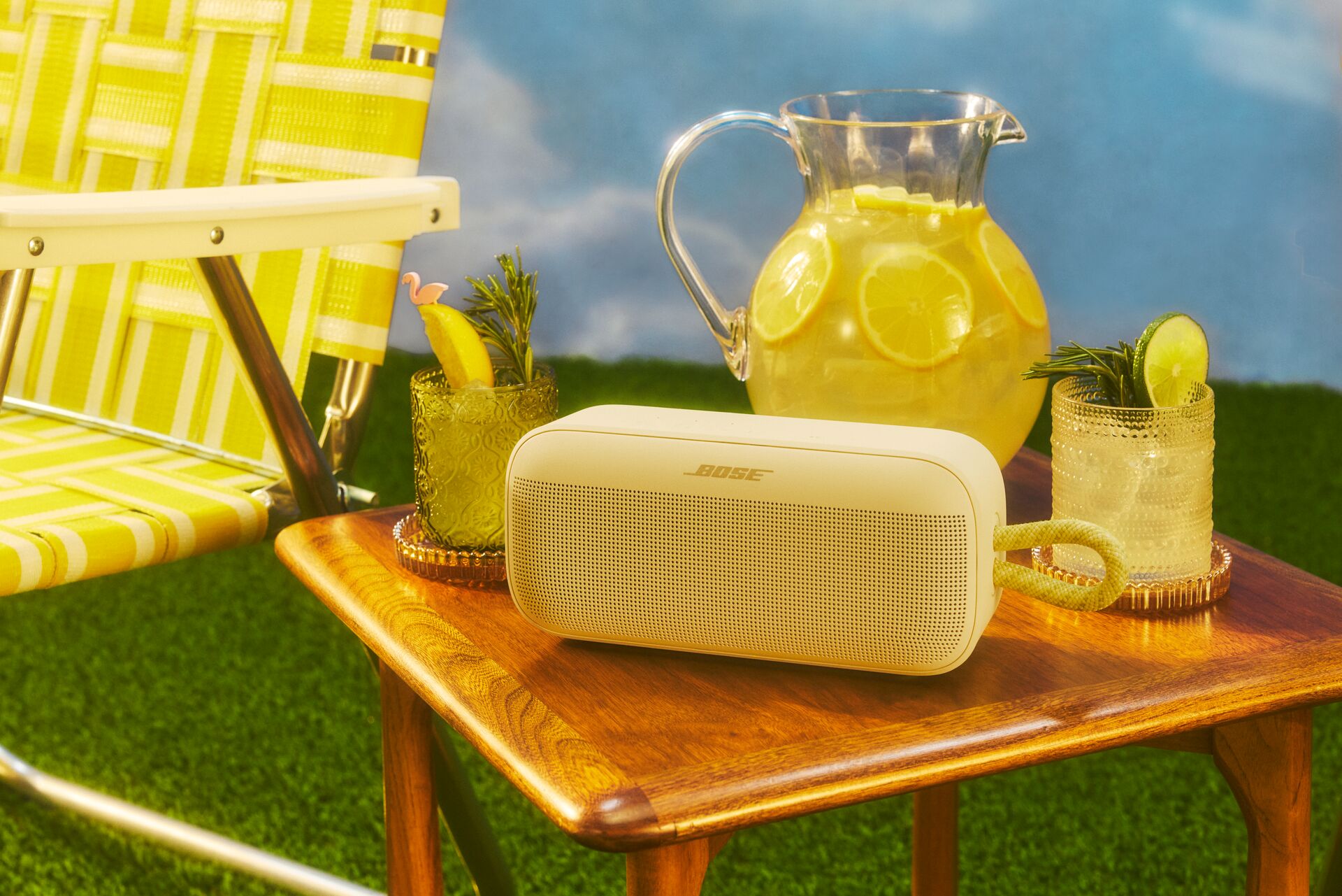 The Bose SoundLink Plus speaker (yellow) sitting next to yellow objects. These include a pitcher of lemonade, yellow glasses, and a yellow lawn chair.