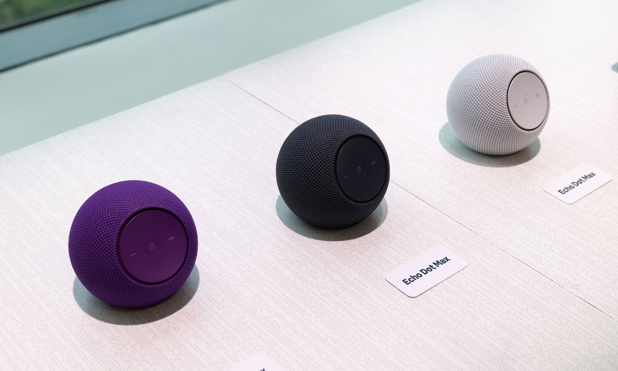 Three Amazon Echo Dot Max speakers, one purple, one white and one black, are displayed on a white wooden table.