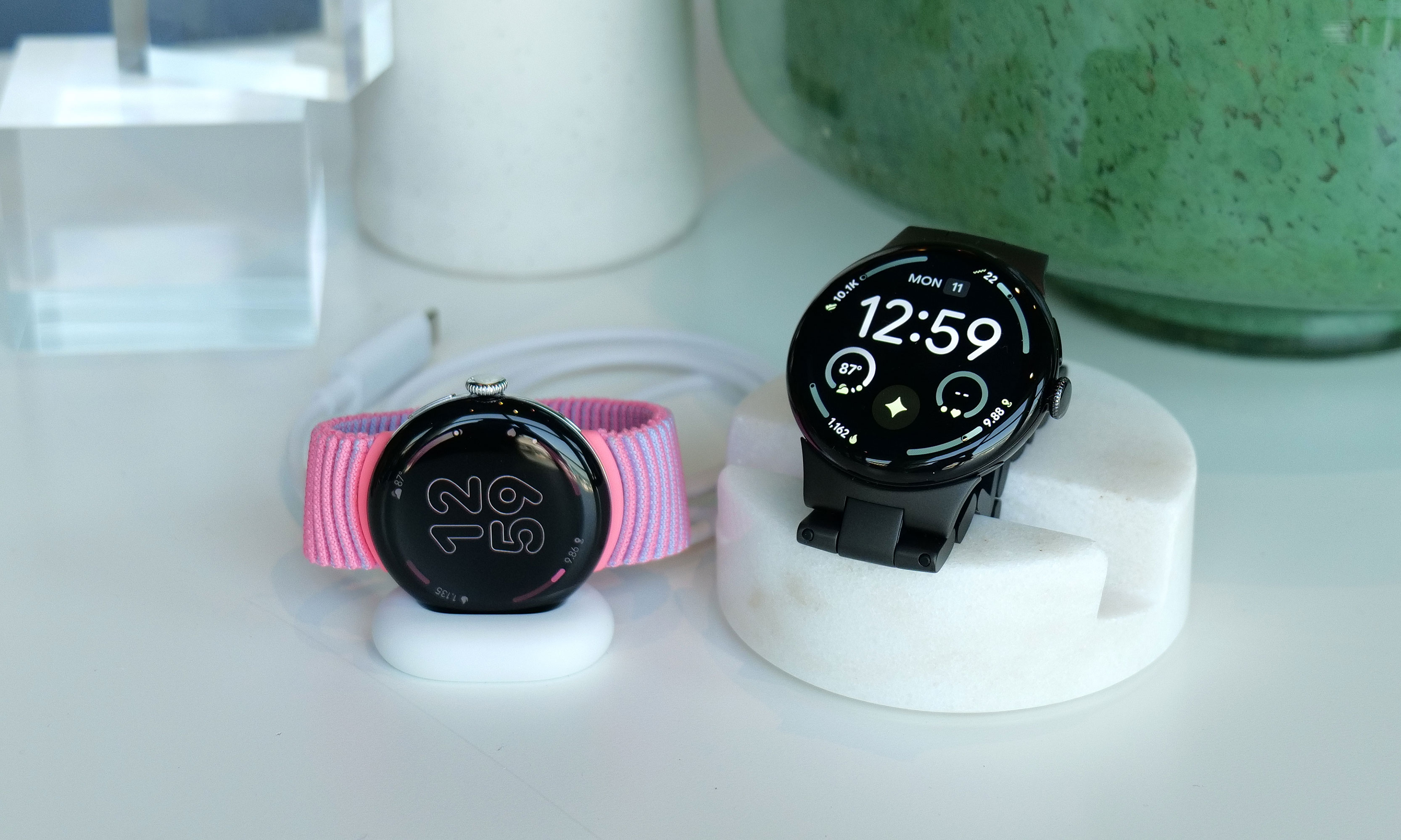 A Pixel Watch 4 laid on its side in its charging cradle, next to a larger Pixel Watch 4 propped up on a faux marble stand.