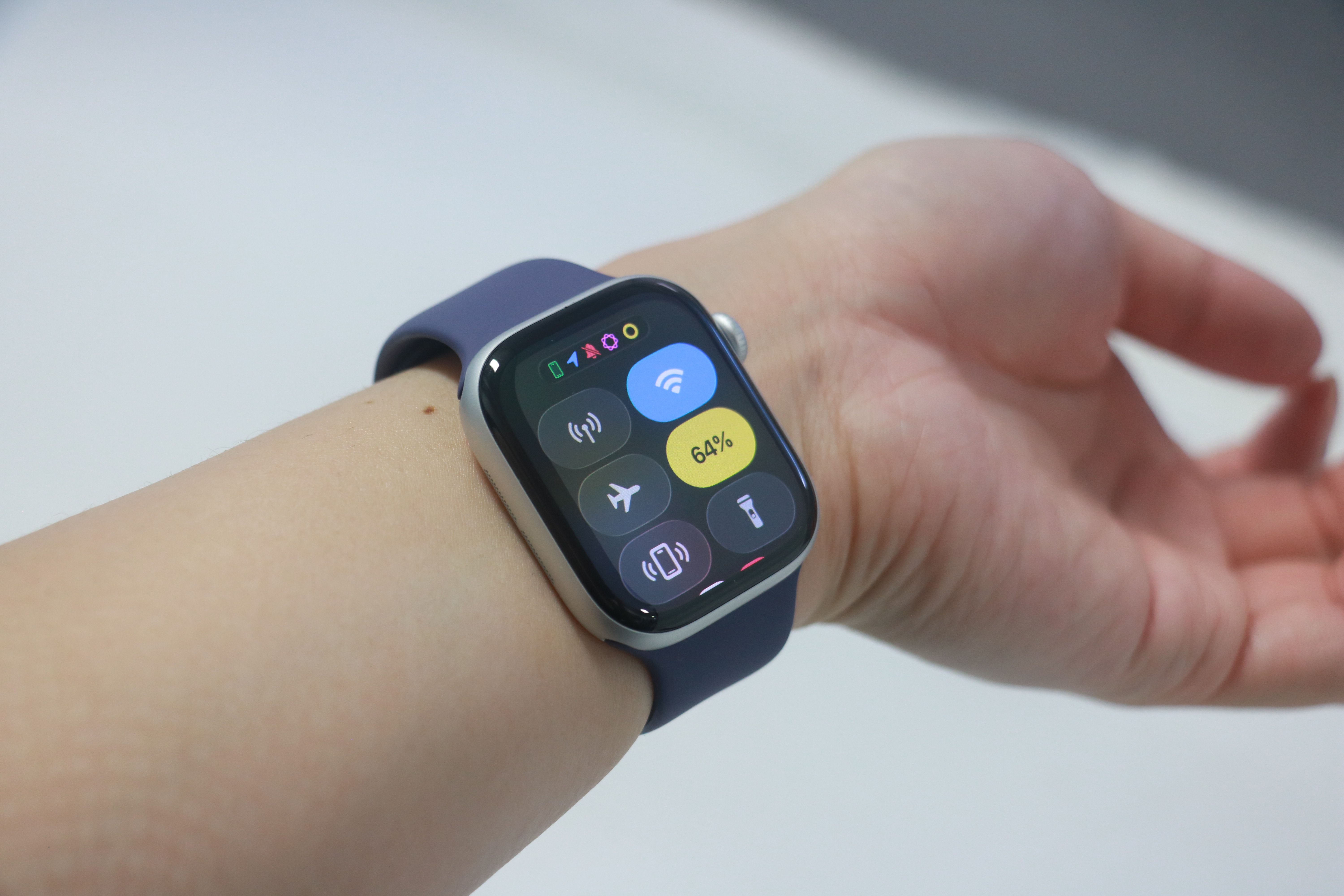 The Apple Watch Series 11 on the inside of a person's wrist, showing the Control Center onscreen. The battery percentage tile is highlighted in yellow, indicating low power mode is on, and the power level is at 64 percent.