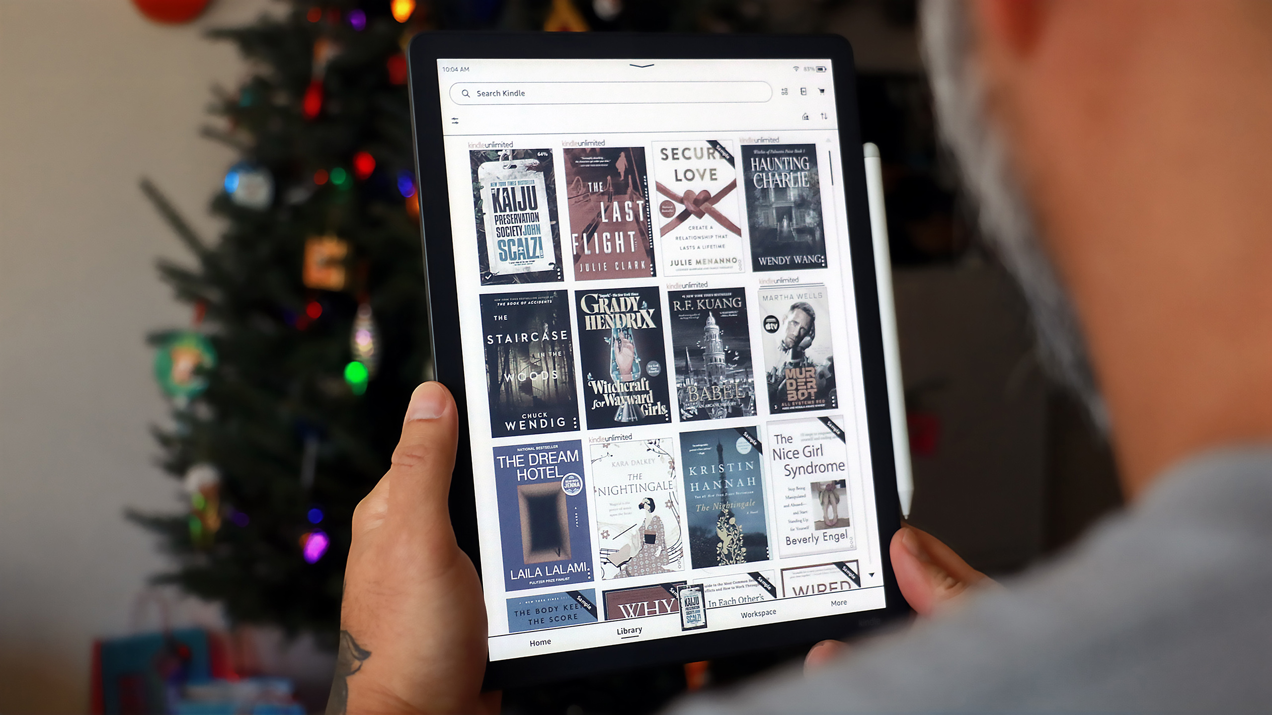 A Kindle Scribe Colorsoft held up in mid-air in front of a Christmas tree, showing a library of book covers in color.