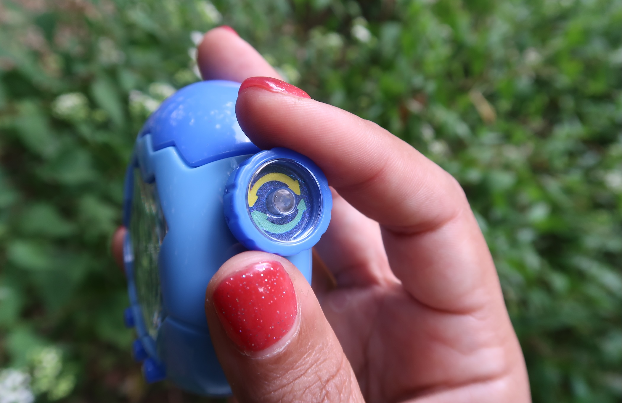 A Tamagotchi Paradise Blue Water device is pictured side-on to show the zoom dial pinched between a person's thumb and index finger