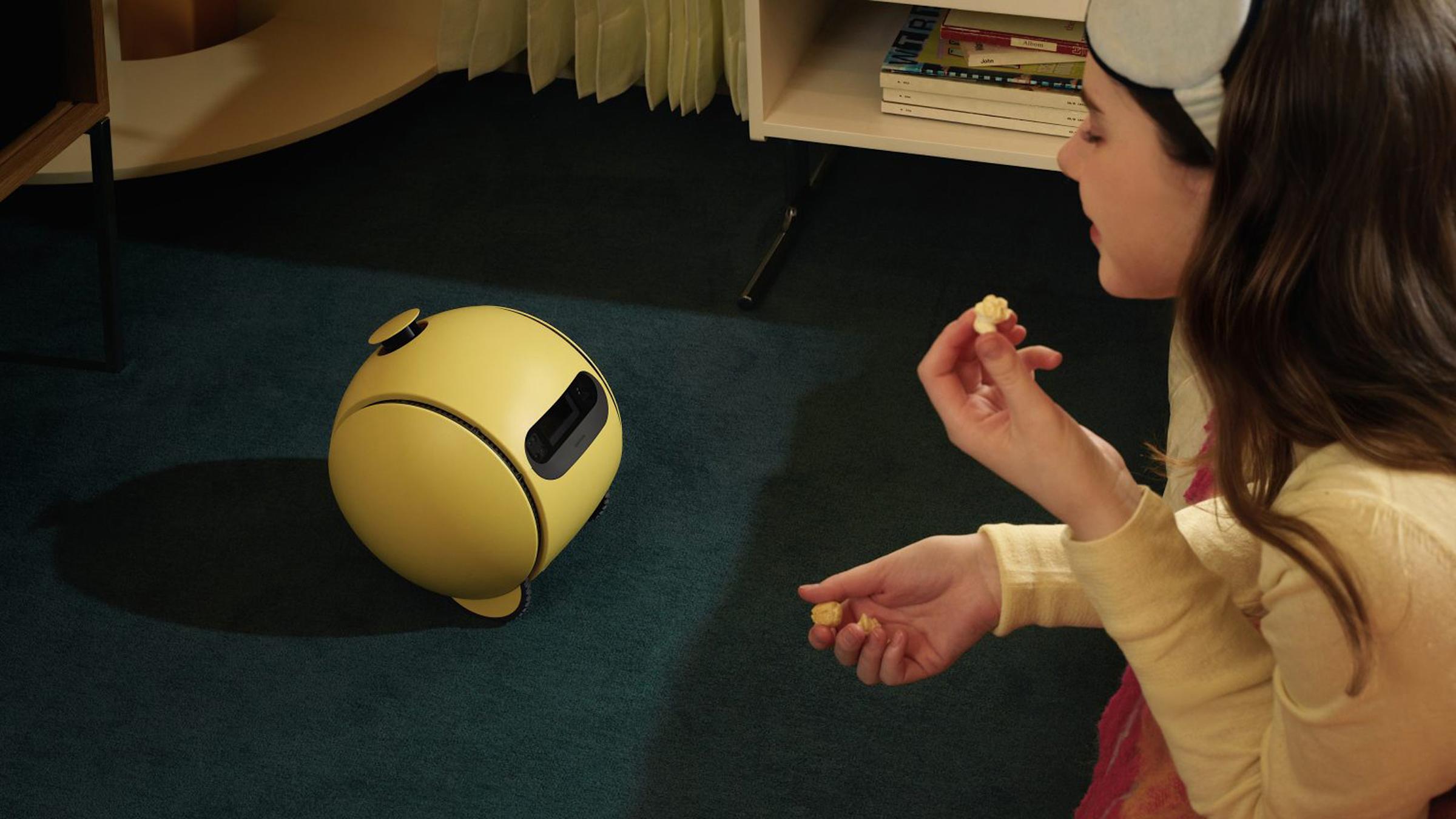 A woman talking to a yellow Samsung Ballie robot on the floor.