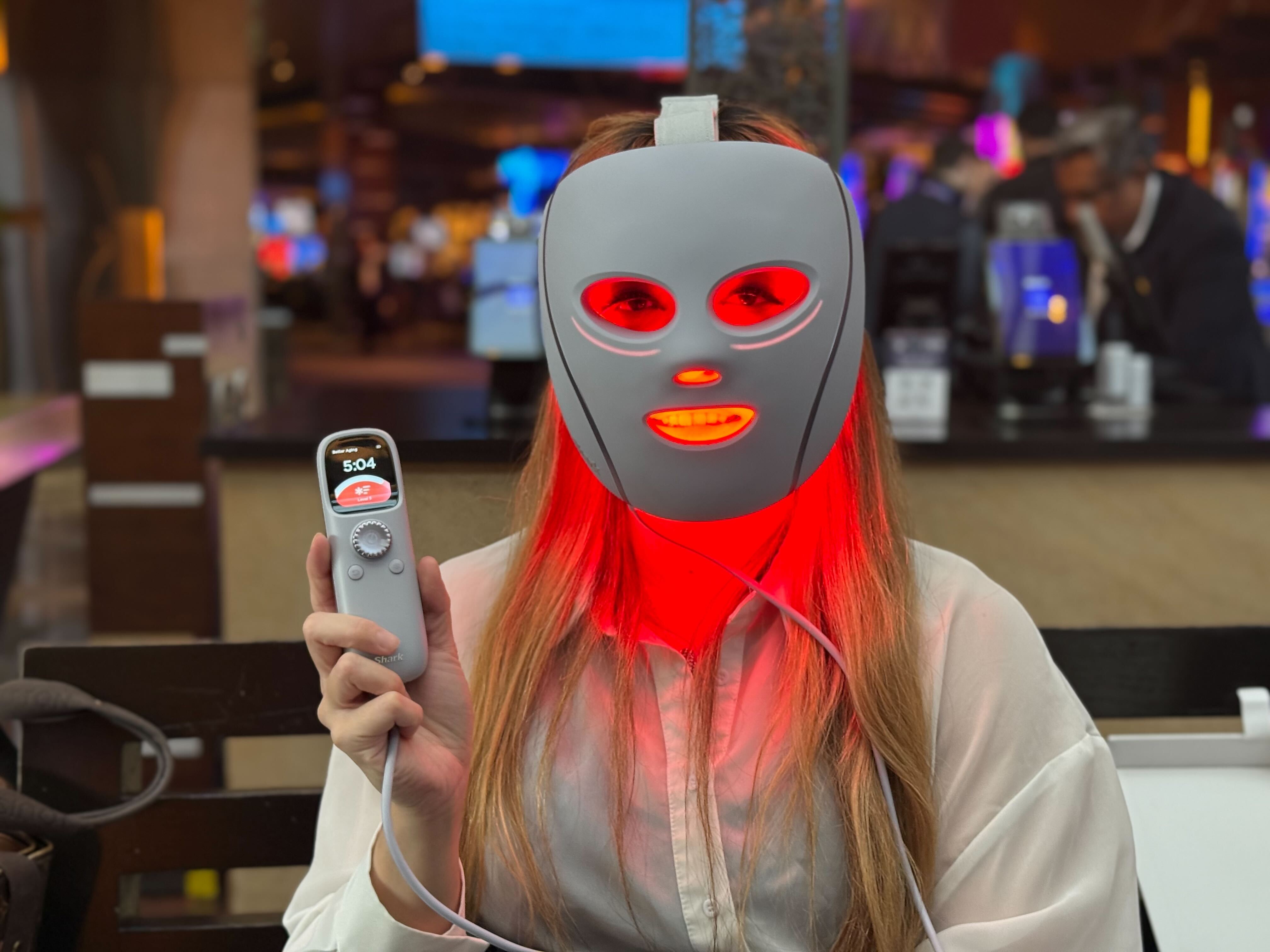A woman wearing the Shark CryoGlow mask, which is glowing with red light out of its holes. The woman is also holding up a remote control with a dial and a small screen.