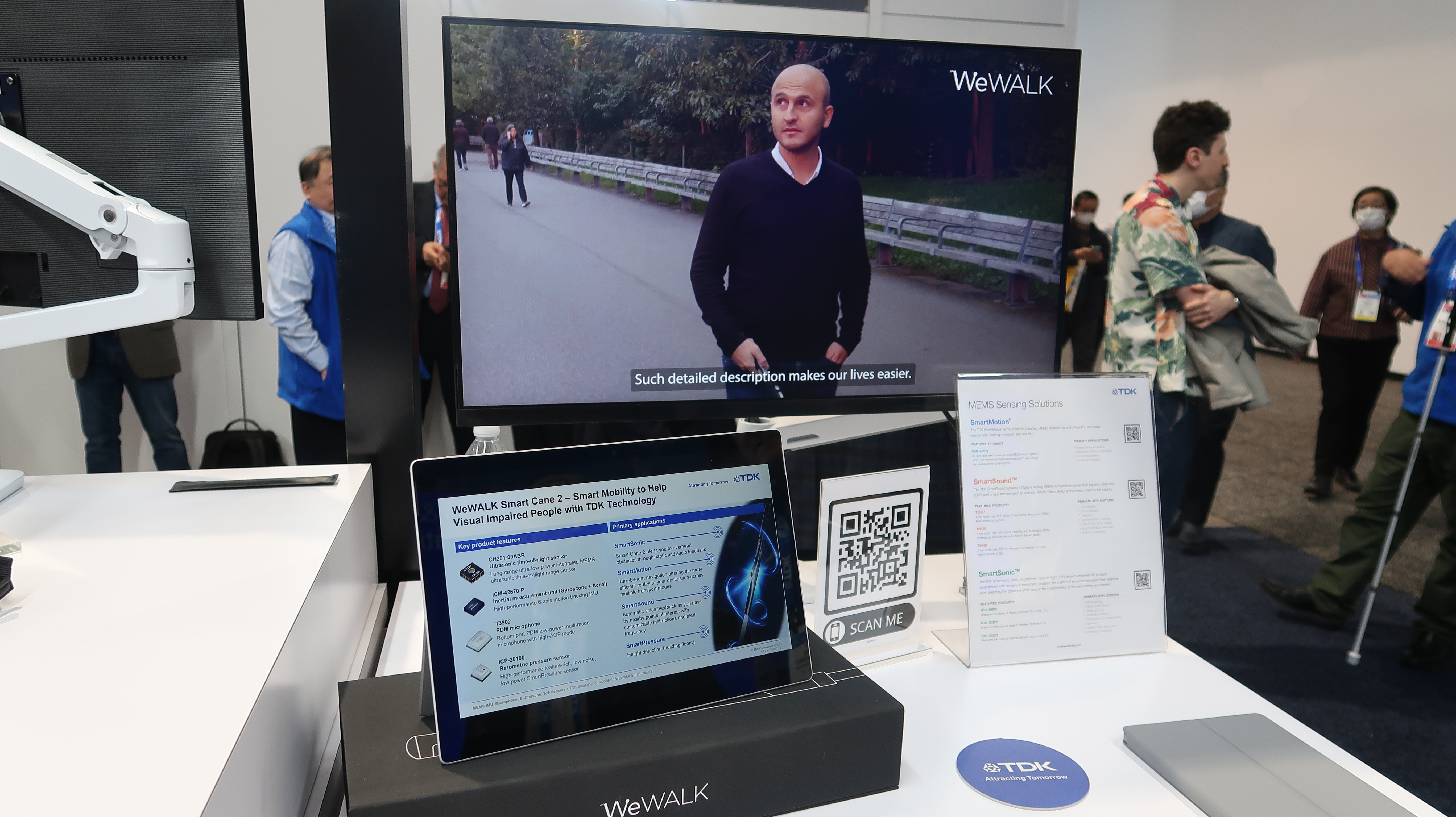 An tablet is positioned on a stand on a white table displaying detailed information about the sensors inside the smart cane. A monitor behind the tablet shows co-founder Kursat Ceylan walking on a path and gripping the handle of the cane. a caption at the bottom says 