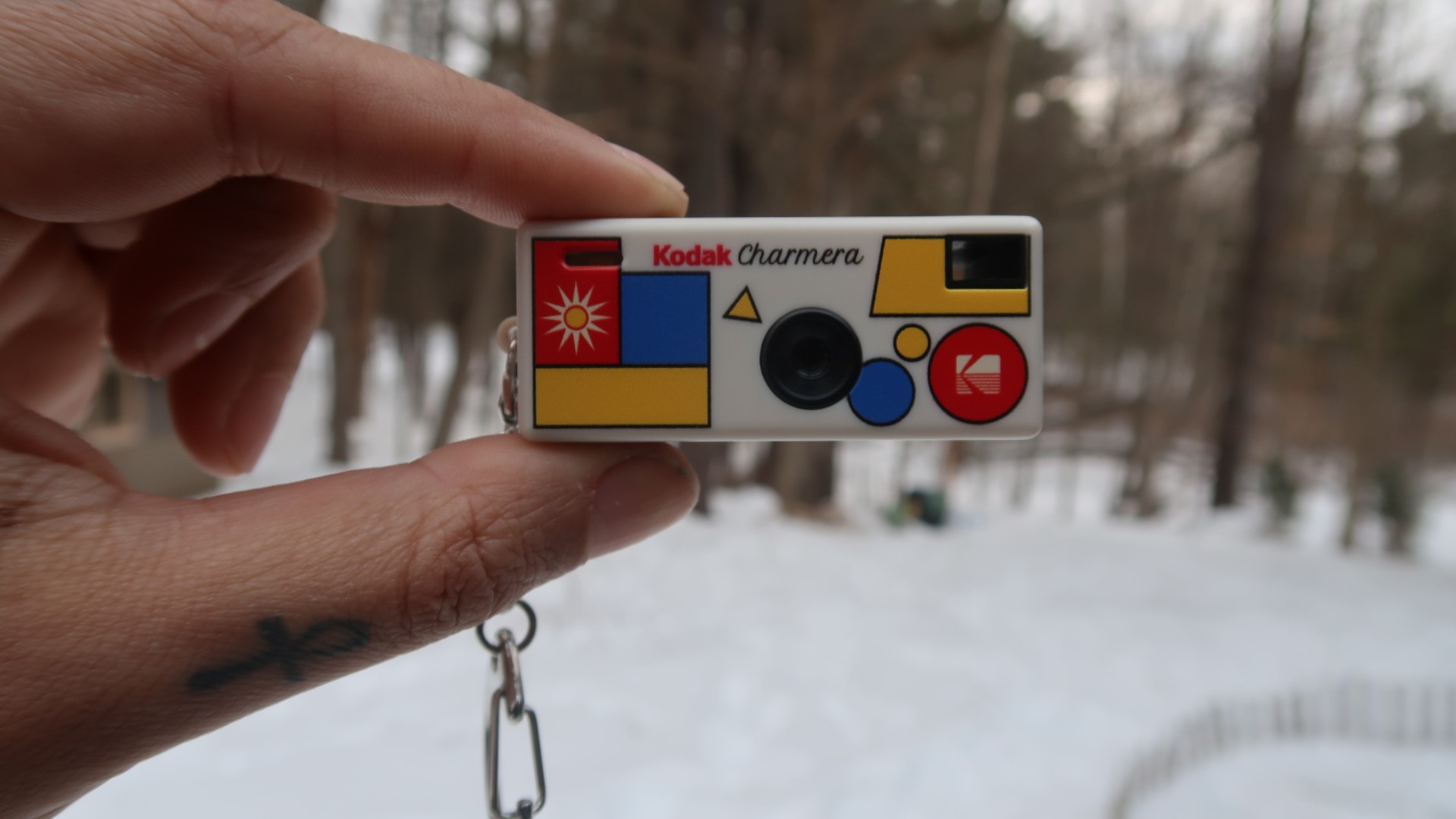A Kodak Charmera is shown pinched between two fingers, with snow and trees seen in the background