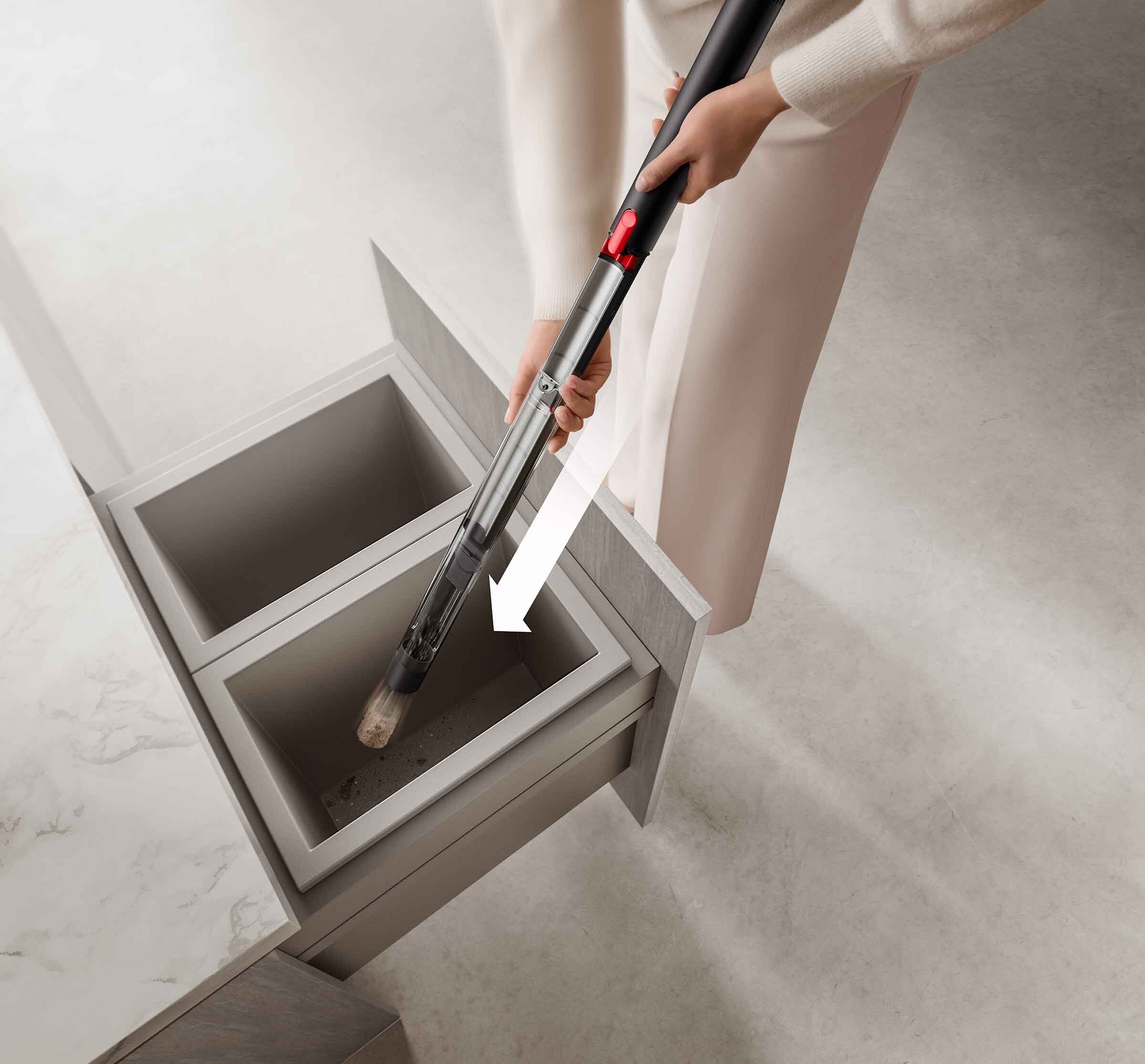 A pair of hands holding the PencilVac over a trash container, with an overlaid arrow indicating downward movement of the vacuum cleaner's bin.