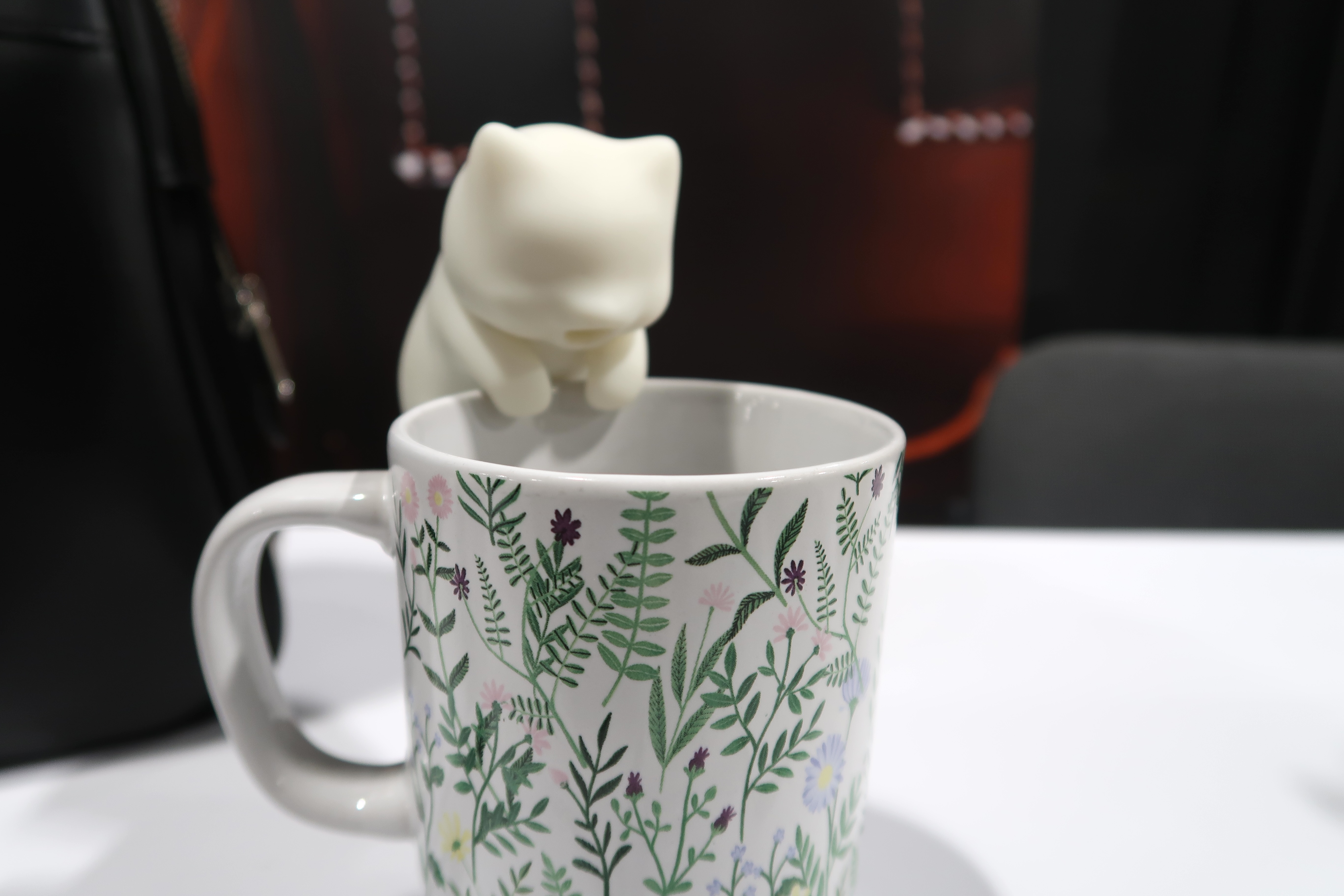 A silicone white cat shaped device is perched on the rim of a mug blowing air onto it
