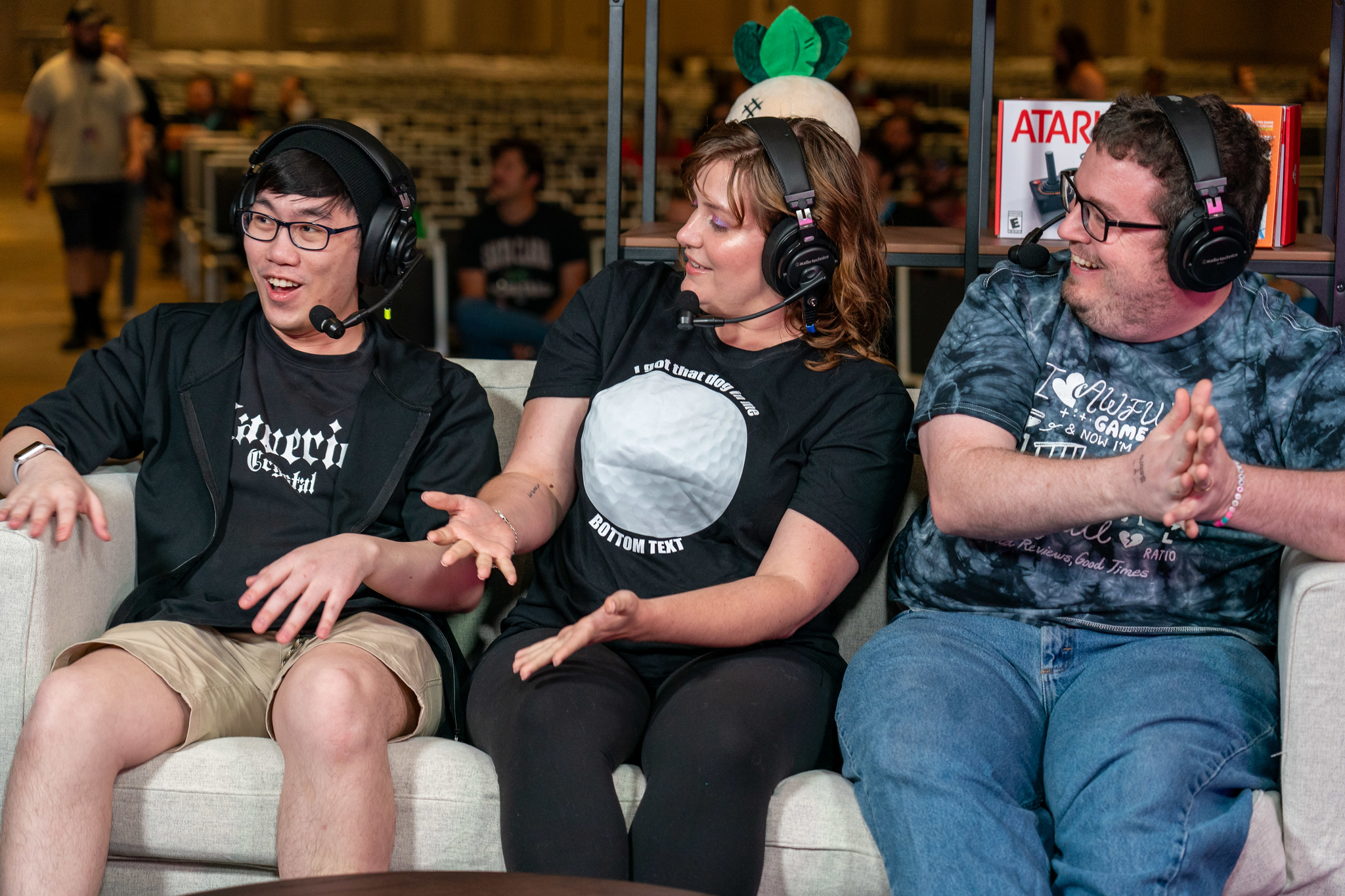 Three gamers on a couch, having fun. Headphones on. Gesturing toward the person on the left.