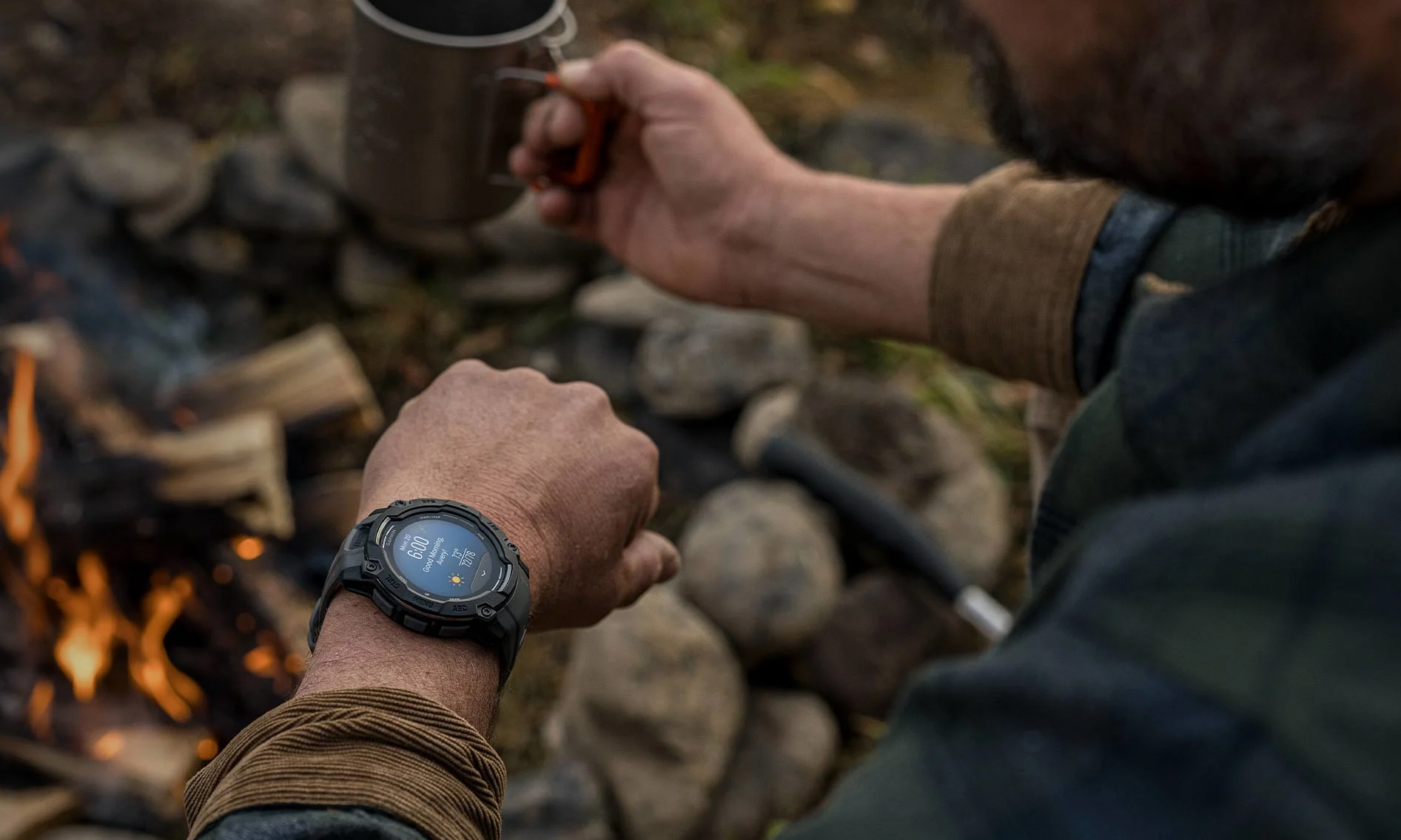 A person sitting by an outdoor fire, wearing a Garmin GPS watch, holding wrist up with coffee in the opposite hand.