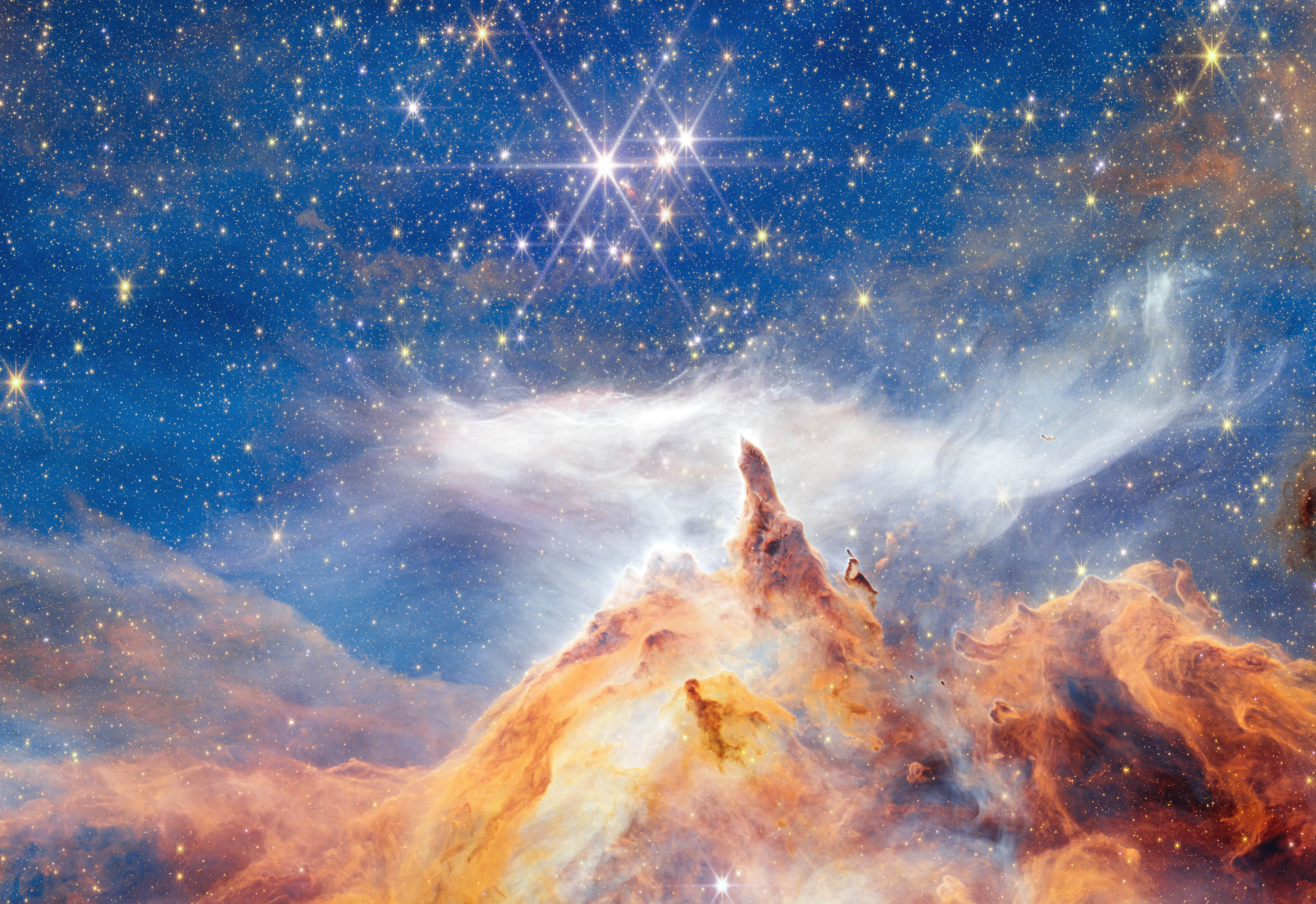 This sparkling scene of star birth was captured by the NASA/ESA/CSA James Webb Space Telescope. What appears to be a craggy, starlit mountaintop kissed by wispy clouds is actually a cosmic dust-scape being eaten away by the blistering winds and radiation of nearby, massive, infant stars. Called Pismis 24, this young star cluster resides in the core of the nearby Lobster Nebula, approximately 5,500 light-years from Earth in the constellation Scorpius. Home to a vibrant stellar nursery and one of the closest sites of massive star birth, Pismis 24 provides rare insight into large and massive stars. This region is one of the best places to explore the properties of hot young stars and how they evolve. [Image description: In what appears as a celestial dreamscape, a blue and black sky filled with brilliant stars covers about two thirds of the image. The stars are different sizes and shades of white, beige, yellow, and light orange. Across the bottom third of the scene is a craggy, mountain-like vista with spire-like peaks and deep, seemingly misty valleys. These so-called mountains appear in varying shades of orange, yellow, and brown. Above their soaring spires is a wispy, ethereal white cloud that stretched horizontally across the scene. Steam appears to rise from the mountaintops and join with this cloud. At the top, right corner of the image, a swath of orange and brown structure cuts diagonally across the sky.]