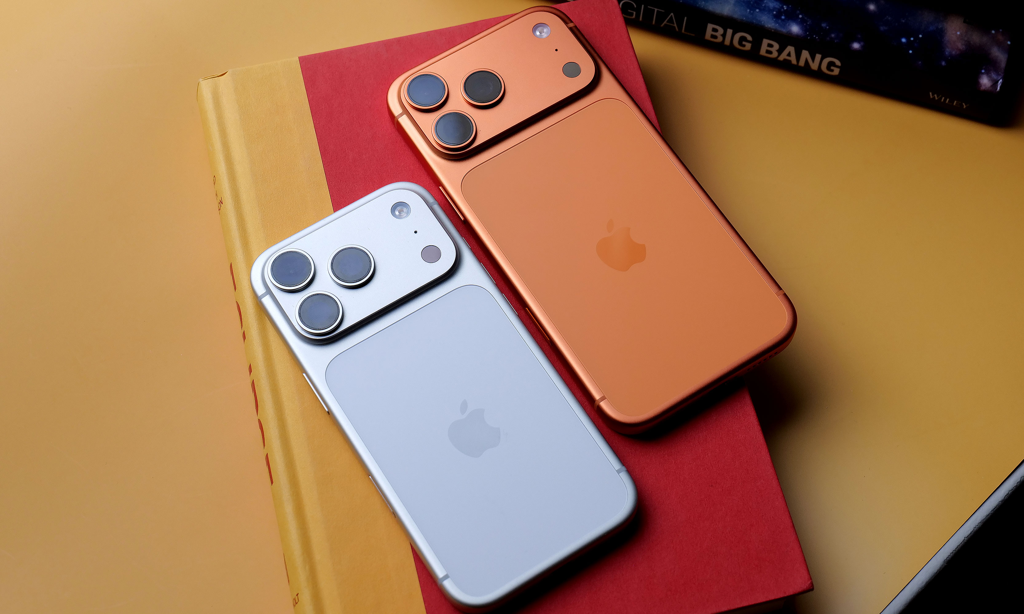 The white iPhone 17 Pro and orange iPhone 17 Pro Max lying face down on a red book.