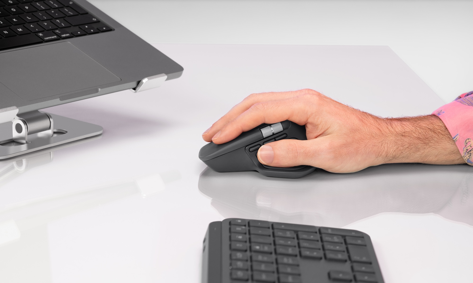 A man uses a graphite Logitech MX Master 4 wireless mouse on a white glossy desk.