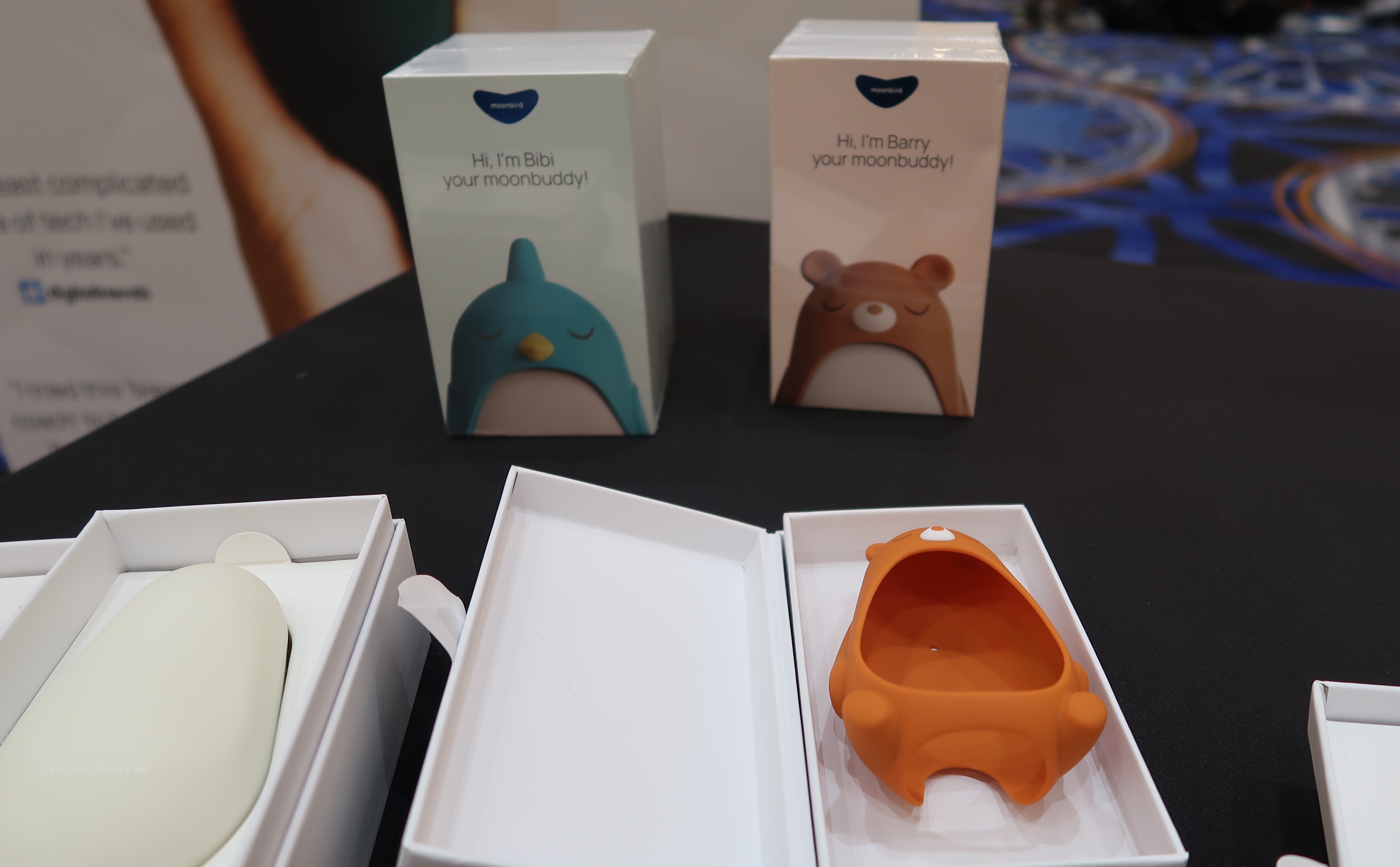 A Moonbuddy device pictured next to an empty Bear sleeve, with two product boxes in the background showing the bird and bear designs