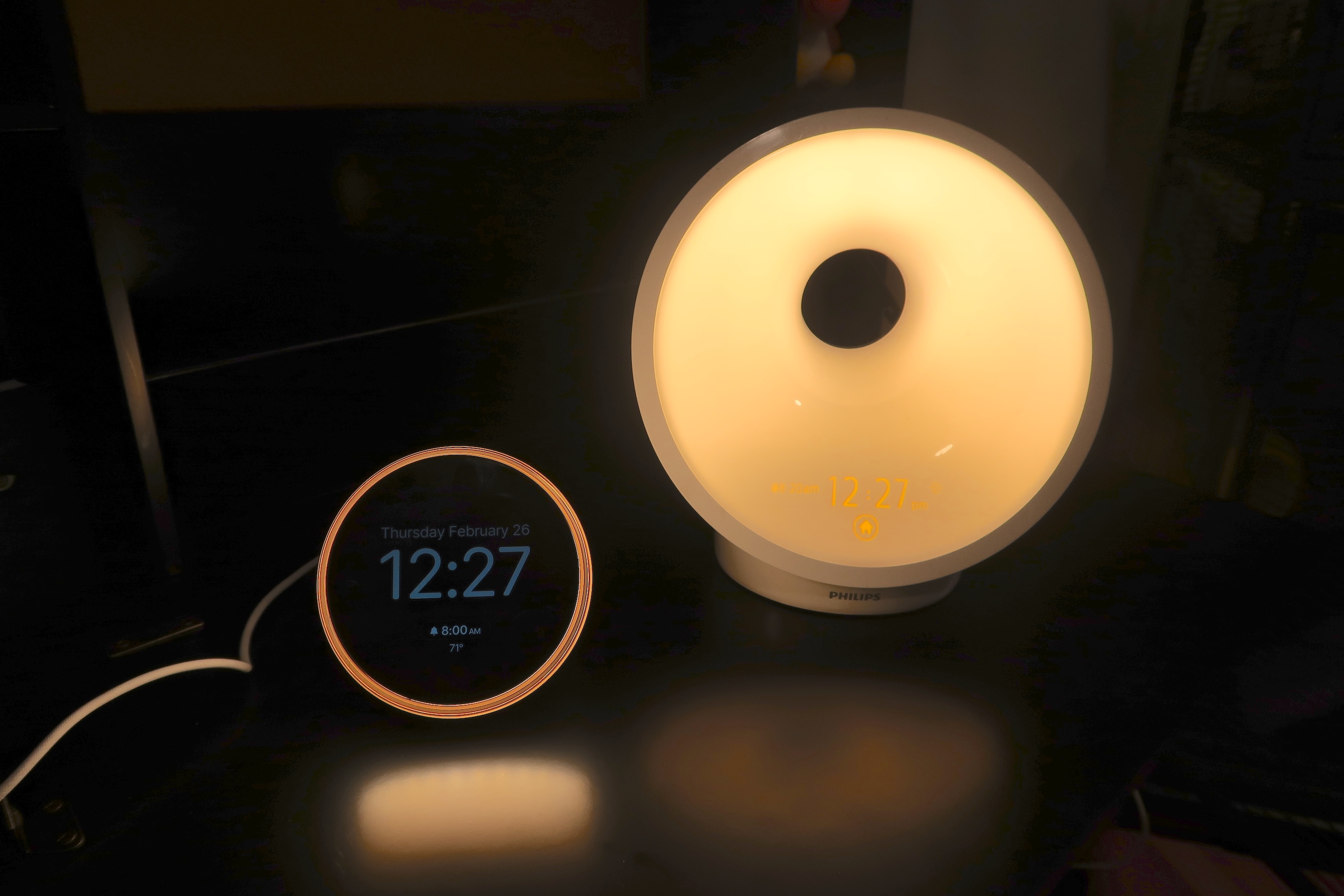 Dreamie alarm clock displaying the time (12:27pm) with a ring of light around the display, beside a much larger Philips Wakeup Light, with the light on and time displayed