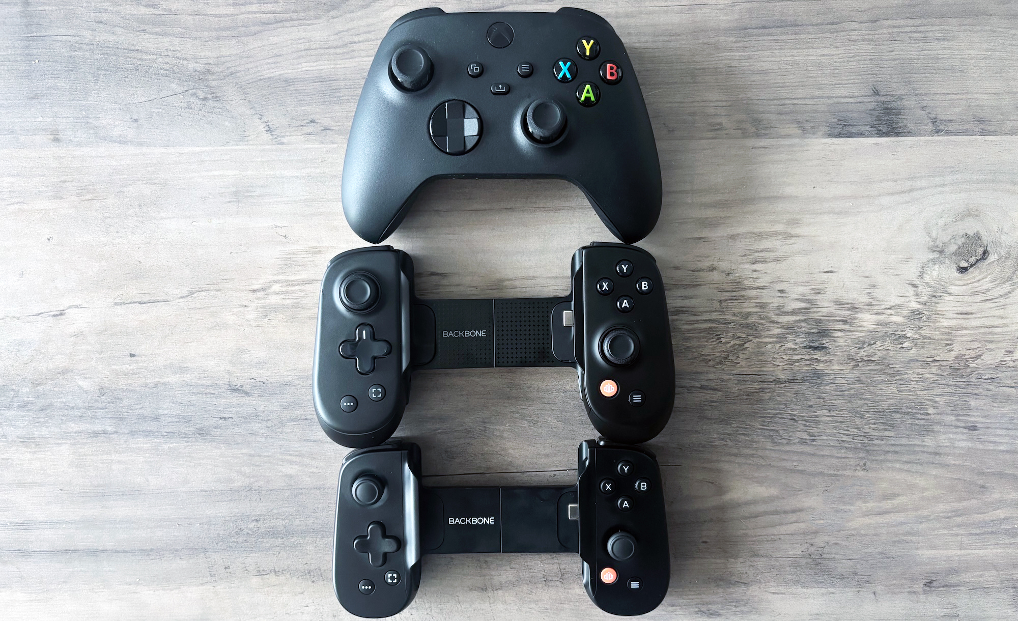 The Backbone Pro mobile game controller sits between an Xbox Series X/S controller and a Backbone One on a light brown wooden table.
