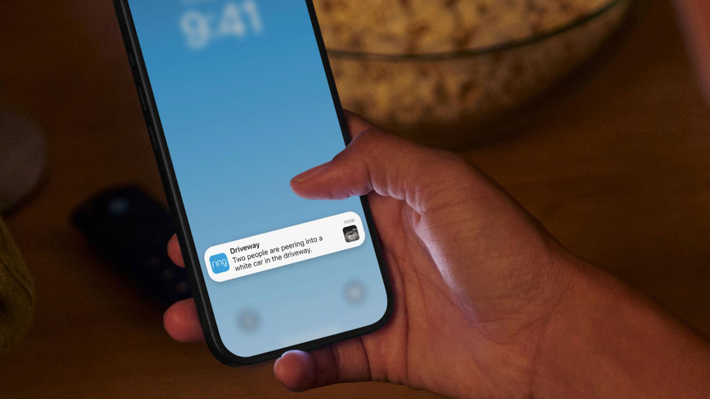 A person's phone with a Ring notification. The alert describes two people peering into a car.