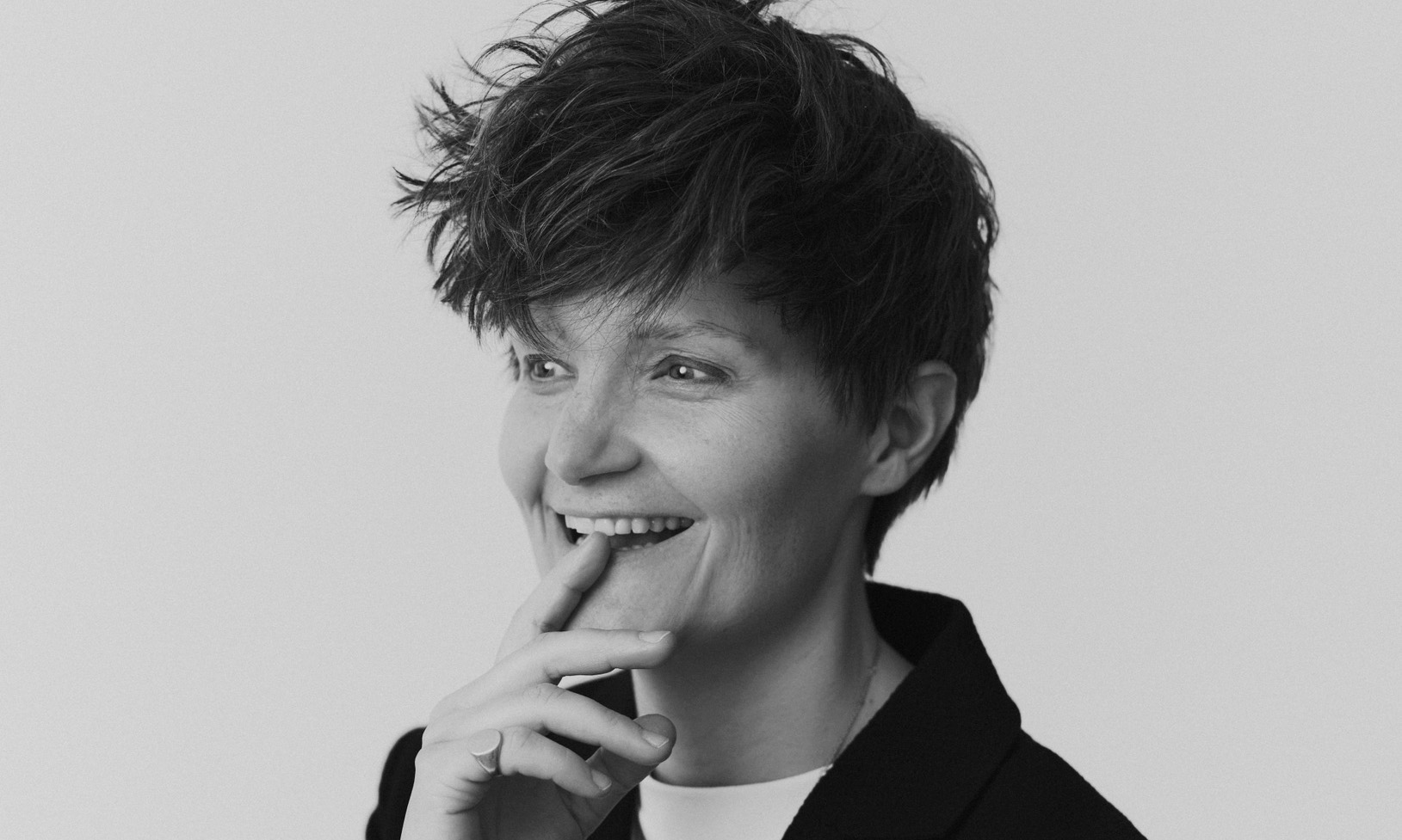 Headshot for writer and actor Charlie Covell. They smile with an index finger pressed to their mouth.