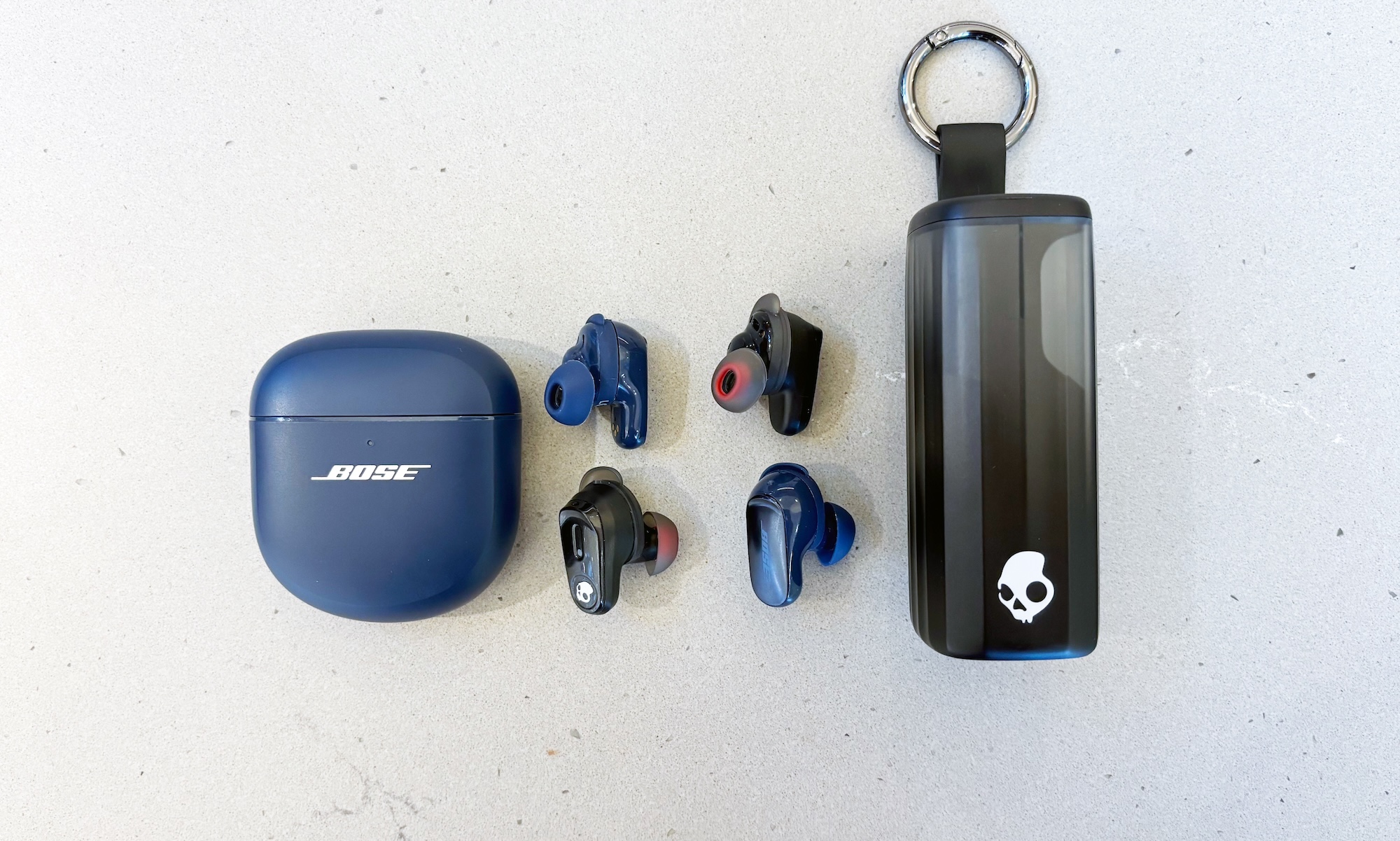 Two pairs of wireless earbuds, the Bose QuietComfort Ultra Earbuds and the Skullcandy Method 360 ANC, sit next to one another on a slate gray countertop. The earbuds are situated between the two pair's charging cases. The Bose pair is navy blue, while the Skullcandy pair is black.