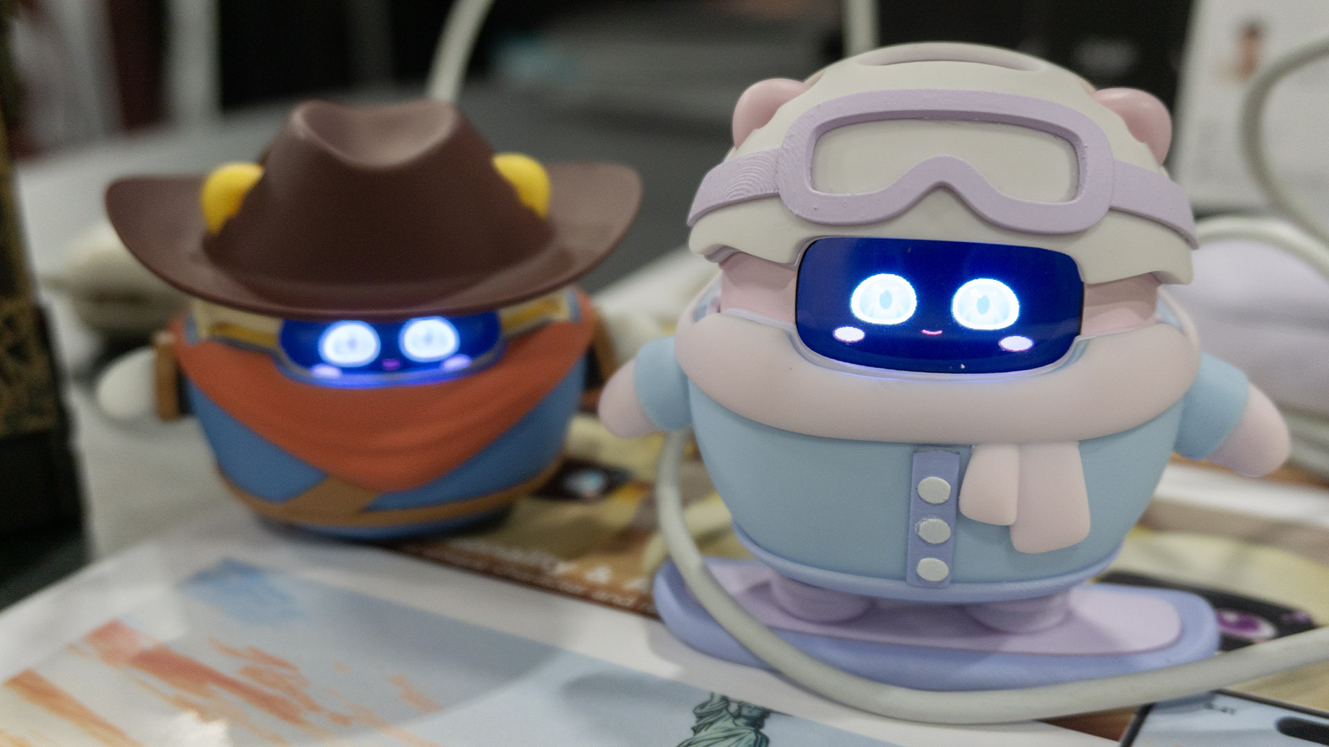 Two Sweekar devices are pictured on a table, one wearing a pink and blue snowboarder outfit and the other (behind it) wearing a cowboy hat and outfit