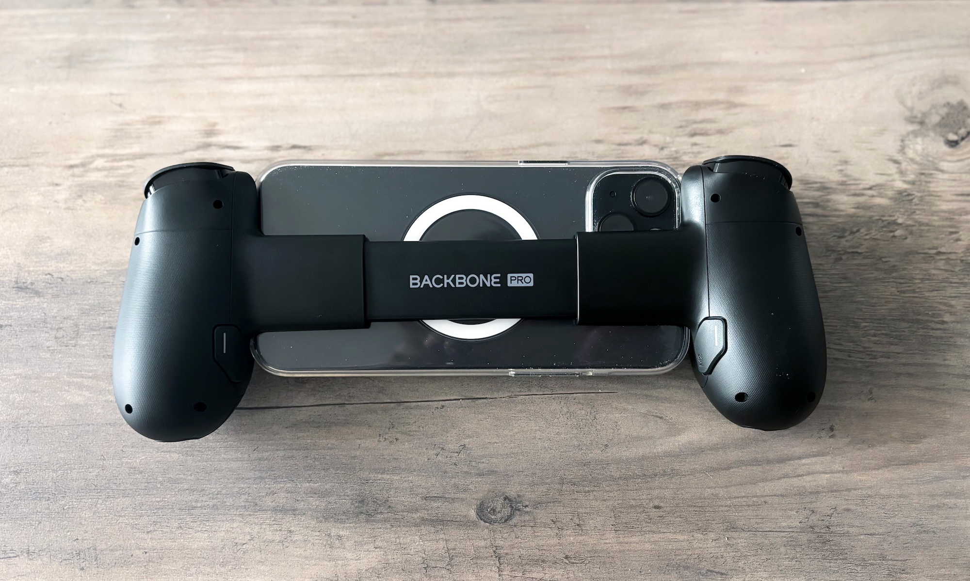 The back of the Backbone Pro mobile game controller is shown connected to an iPhone 15 Plus on a light brown wooden table.