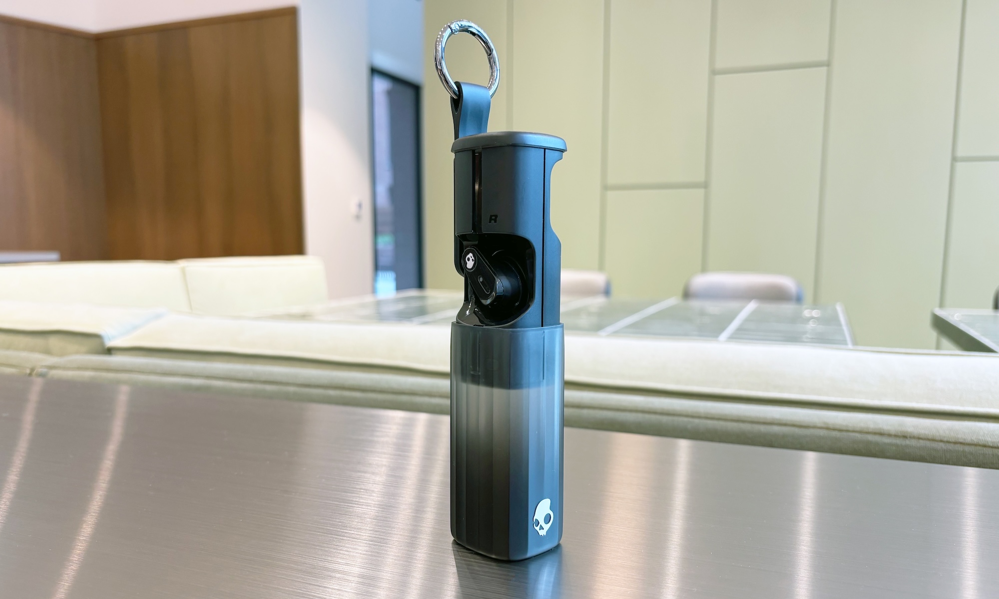 A pair of black wireless earbuds, the Skullcandy Method 360 ANC, sit inside their tube-like charging case on a silver desk in front of a pale green wall. The charging case is extended and slid open to show how the earbuds fit into the case.
