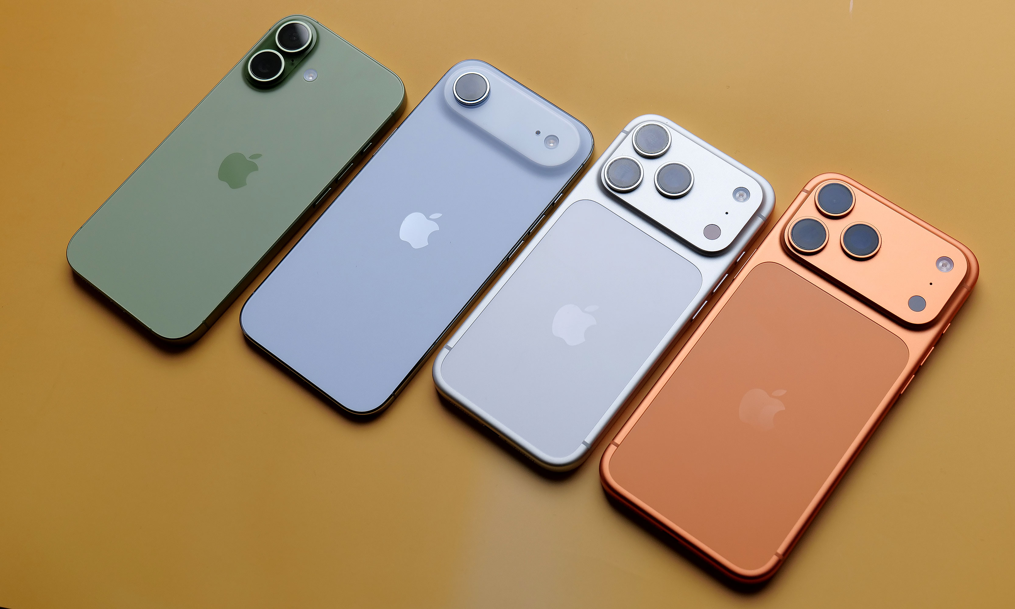 The iPhone 17, iPhone Air, iPhone 17 Pro and iPhone 17 Pro Max laid out face down in a row on a dark yellow surface.